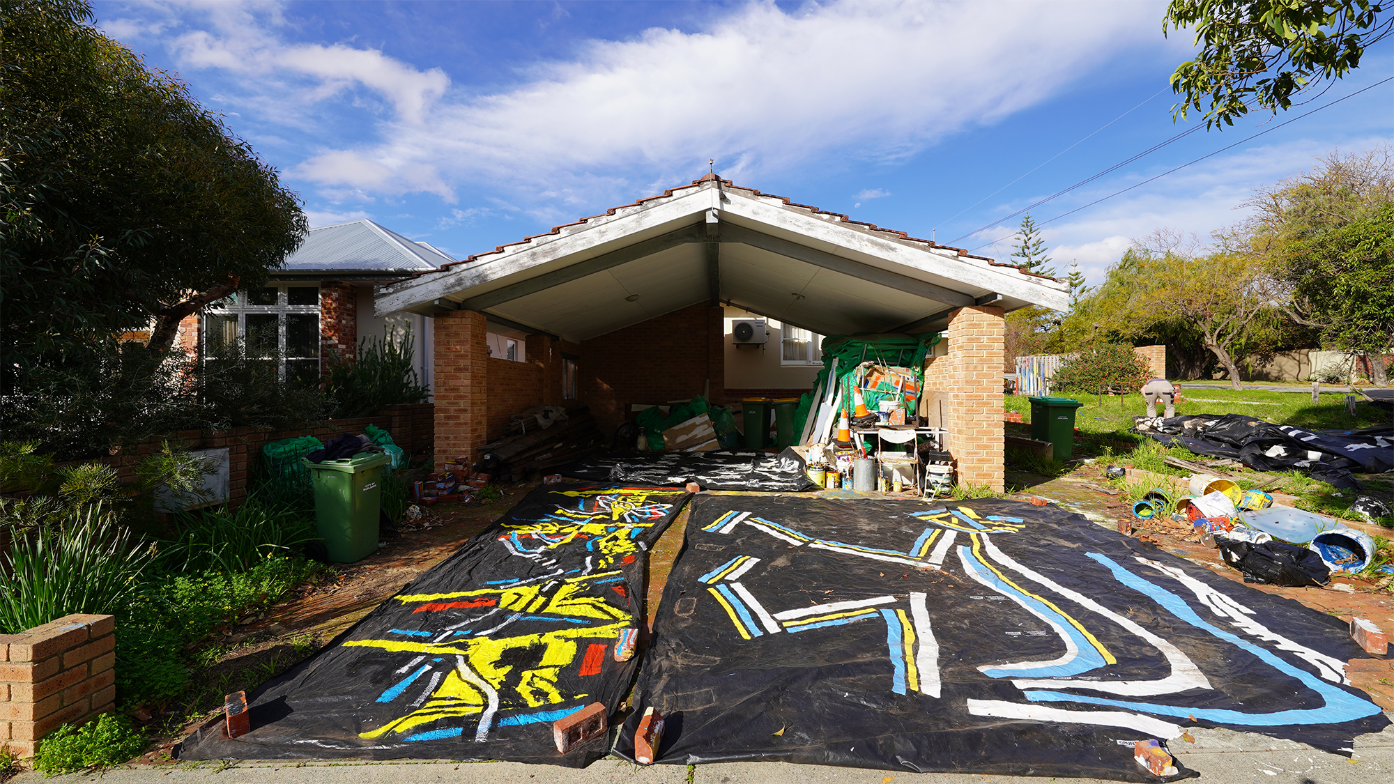 Photograph of a garage with two large plastic paintings spread out on the driveway and an array of paint materials and supplies scattered around